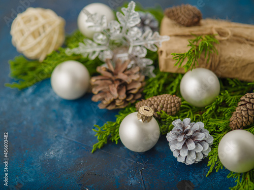 Stylish composition of fir branches, cones, white Christmas balls and an original wrapped gift on a blue background. New Year, Christmas, postcard, invitation, banner, poster.