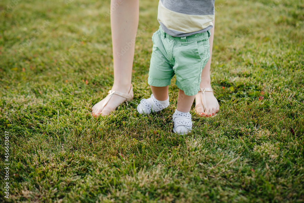 A young cute mother helps and teaches her little son to take his first steps during sunset in the park on the grass. A young cute mother helps and teaches her little son to take his first steps during sunset in the park on the grass.