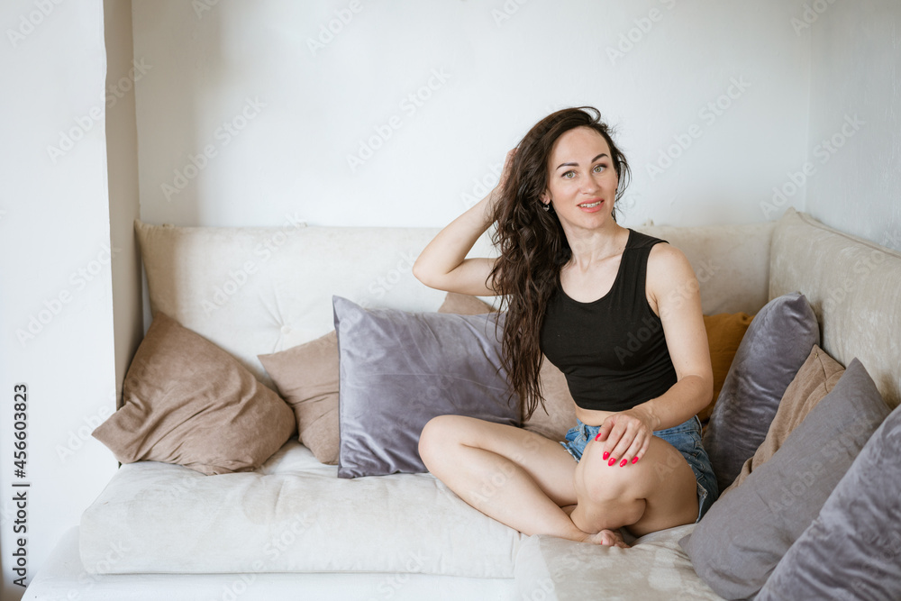a pretty young brunette woman in a black top and blue denim shorts is sitting on the sofa, her legs tucked under her, among many pillows a pretty young brunette woman in a black top and blue denim shorts is sitting on the sofa, her legs tucked under her, among many pillows