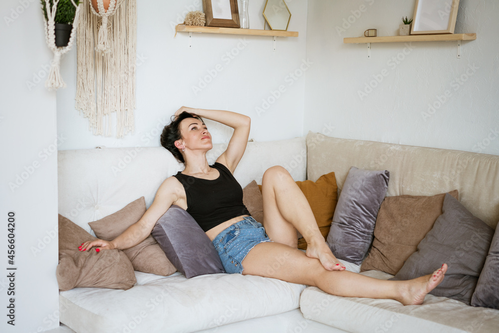a pretty young brunette in a black top and blue denim shorts is lying on the sofa among many pillows