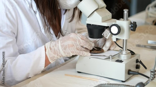 Conservation of archaeological finds in the laboratory. An archaeologist in protective white gloves under a microscope cleans a metal object from Roman times. Processing of archaeological objects
