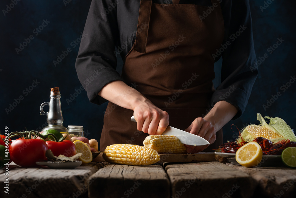 The chef cuts the corn into pieces on a cutting board. Next on the table are ingredients for a traditional American dish - boiled crawfish and corn with vegetables and lemon. Cookbook, restaurant.
