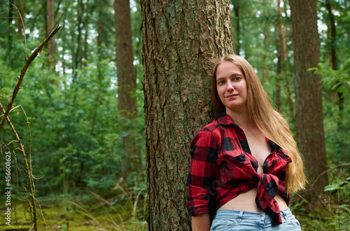 A young Italian woman wearing a red flannel with jeans and leaning on a tree in a forest