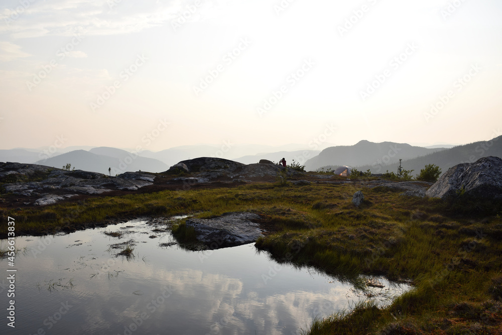 Mont des Morios, Quebec, Canada: View of hiker and camper at the summit during sunset with mountains, pond reflection, alpine vegetation and pitched tent
