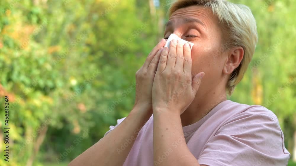 A blonde girl with a short haircut close-up sneezes and blows her nose in a paper napkin against the background of green nature A blonde girl with a short haircut close-up sneezes and blows her nose in a paper napkin against the background of green nature