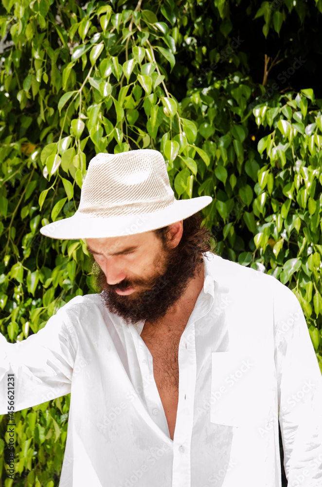 young man with beard and long hair wears hat and white shirt surrounded by greenery