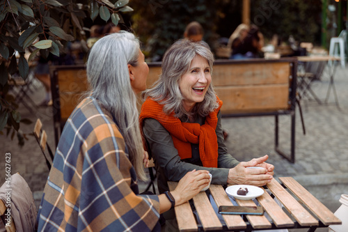 Senior lady laughs spending time with female friend at small table with candies on plate in street cafe on nice autumn day