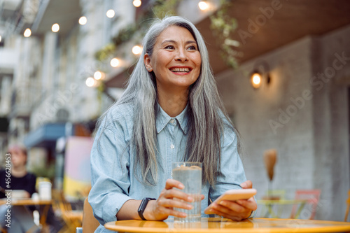 Obraz na plátně Cheerful mature Asian woman with glass of water and mobile phone sits at small t