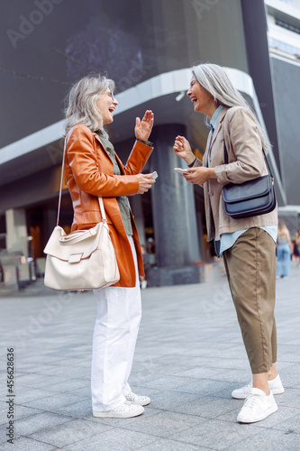 Joyful silver haired senior women with modern mobile phones laugh standing on city street on nice autumn day. Old friends meeting