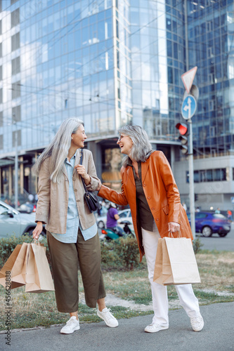 Positive mature woman with shopping bags touches Asian lady elbow laughing on modern city street on autumn day. Friends spend time together