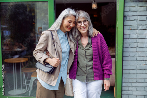 Pretty senior Asian lady hugs grey haired friend in purple jacket standing near cafe entrence on city street. Long-time friendship relationship © Yaroslav Astakhov