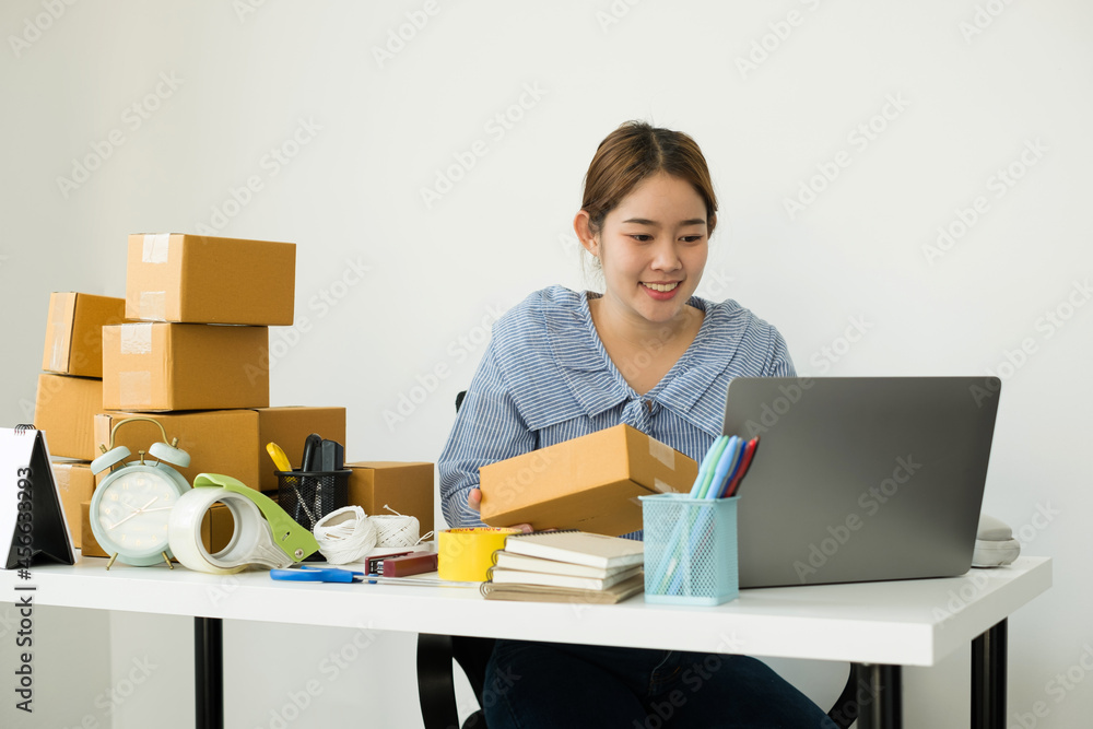 Young Asian woman business owner with many parcel boxes on the table happy online sales job, use the laptop, get an order from customers, take notes, and make arrangements for delivery by post Young Asian woman business owner with many parcel boxes on the table happy online sales job, use the laptop, get an order from customers, take notes, and make arrangements for delivery by post