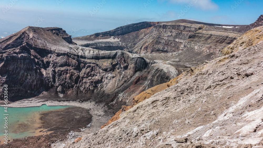 The caldera of an active volcano. The structure of the steep slopes of the crater is visible. At the bottom is a turquoise acid lake with melted snow on the banks. Blue sky. Kamchatka. Uzon