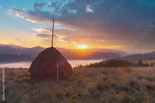 Magnificent sunrise over mountain foggy valley. Panoramic landscape with the sun, through the clouds, rising over Carpathian mountains. Haystack on grassland hill in the foreground.