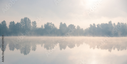 Beautiful foggy morning. Lake coast trees symmetrically reflect in the river water. Fog over autumn lake water.