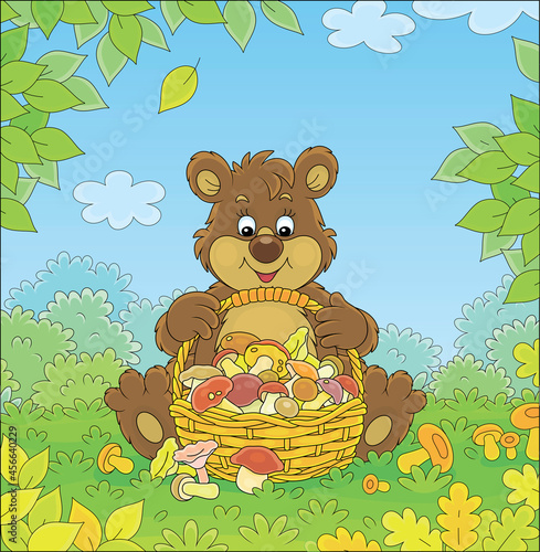 Little brown bear mushroomer friendly smiling and sitting with a big wicker basket full of picked mushrooms on an autumn forest glade, vector cartoon illustration