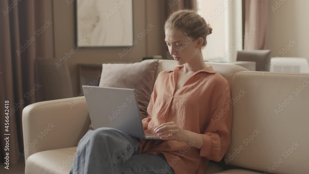 Medium shot of young Caucasian woman in casualwear typing on laptop sitting on cozy sofa in modern living room Medium shot of young Caucasian woman in casualwear typing on laptop sitting on cozy sofa in modern living room
