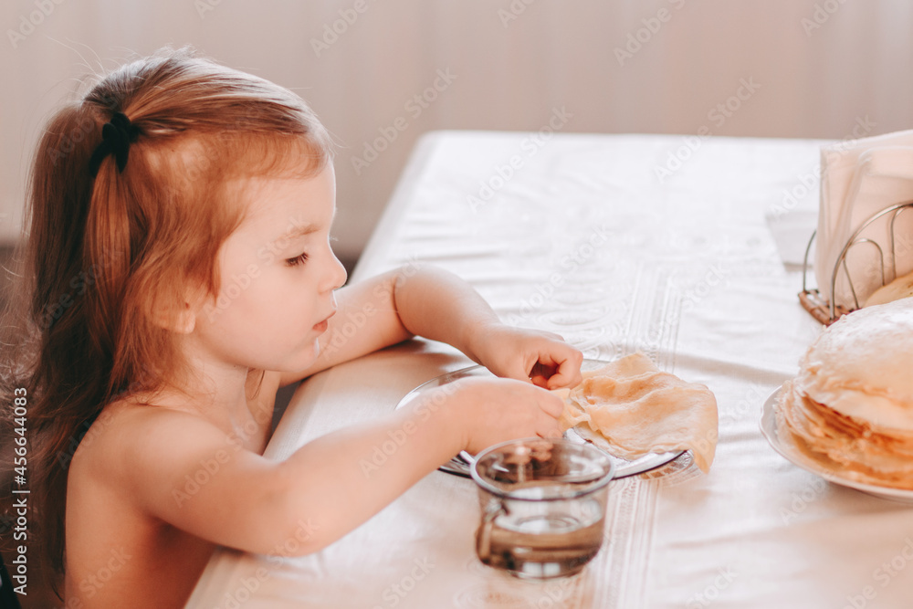 Little girl sitting at table in kitchen and eating thin pancakes crepes.