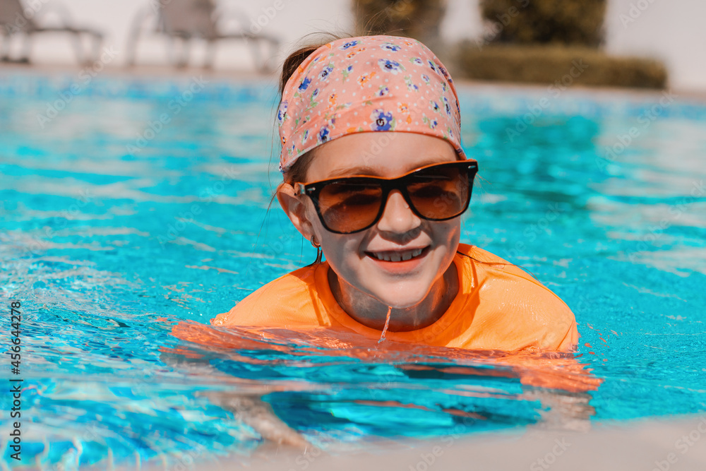 Preteen girl wearing sunglasses swimming in pool with blue water. Healthy and safe summer outdoor activities. Summer tropical vacation in resort.