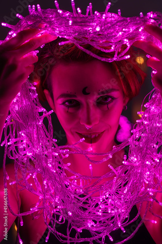 Woman with witch makeup and an earring in her nose. The girl holds a garland of flickering pink lights near her face. Light bulbs illuminate the witch s face in the dark.