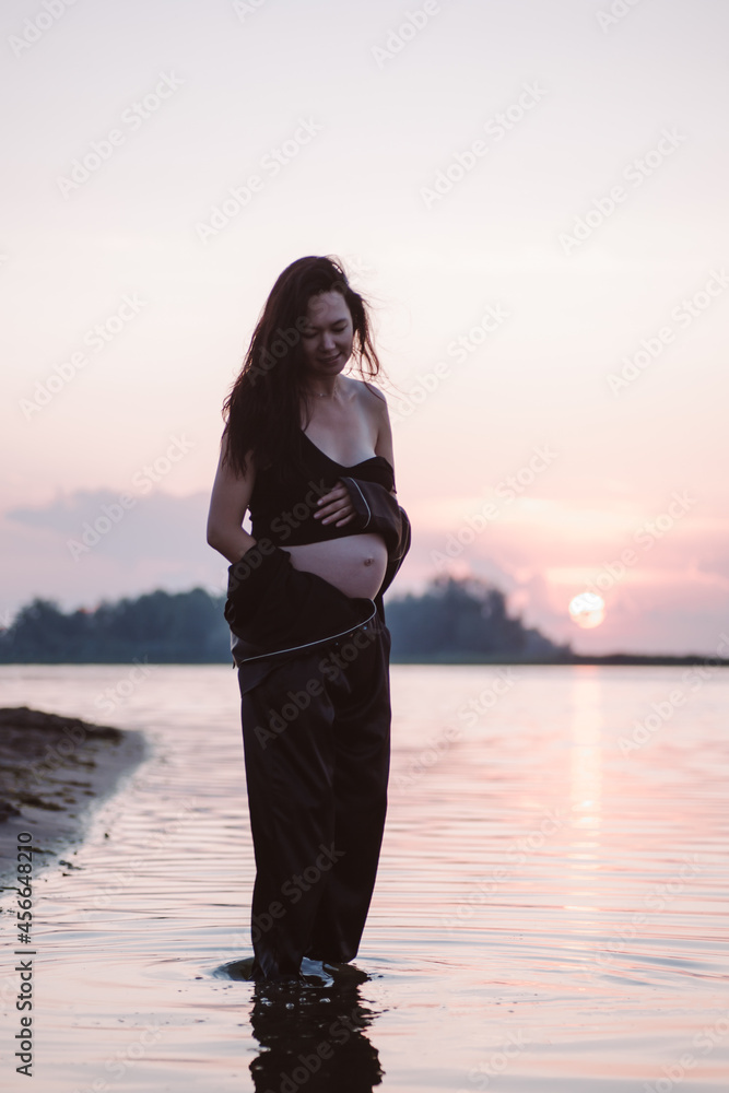 Pregnant woman smiles. Beautiful young pregnant woman in dark suit and long brown hair stands on seashore in rays of setting sun and holds her stomach with her hands. 