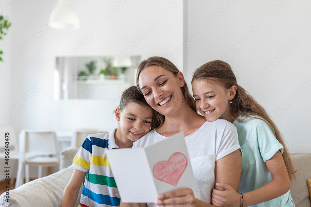 Joyful young mother and little children sitting on sofa and embracing while reading wishes and congratulations with holiday in presented postcard at home