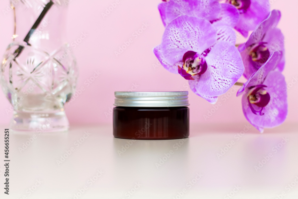 A brown jar of cream on a pink background with beautiful orchids. Stylish appearance of the product, layout, personality. A brown jar of cream on a pink background with beautiful orchids. Stylish appearance of the product, layout, personality.