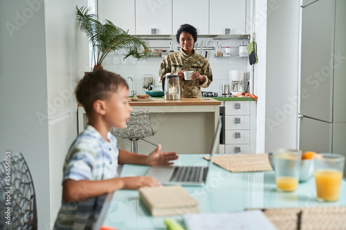 Boy sitting at the table and using laptop while his mother preparing breakfast