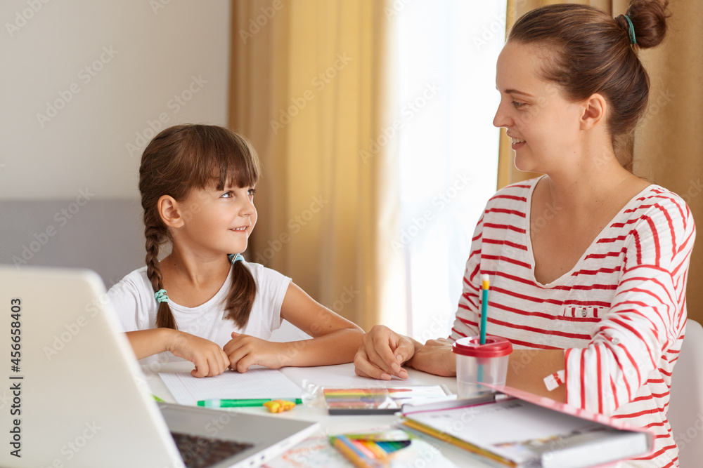 Positive woman with her female kid posing in living room at table, mother helping daughter with lessons, explaining new rule, online distance education. Positive woman with her female kid posing in living room at table, mother helping daughter with lessons, explaining new rule, online distance education.
