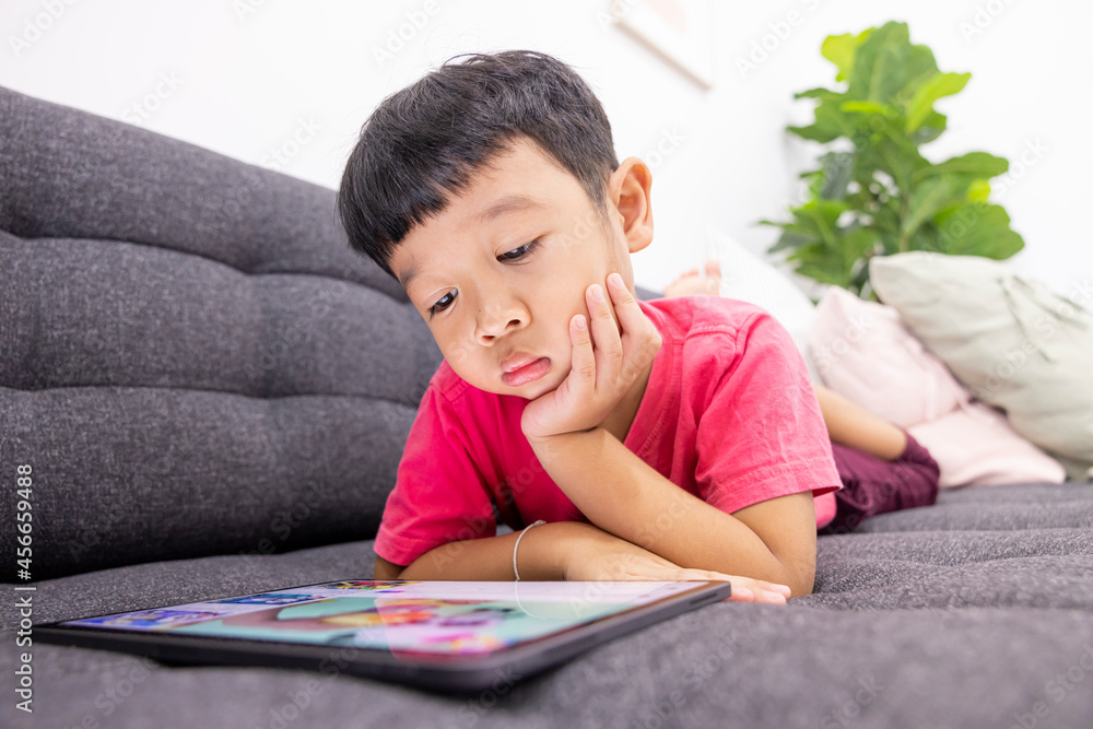 Smiling little boy looks at the touchpad lying on comfortable sofa in the living room. Enjoying playing game on digital tablet. Happy small kid using funny applications Smiling little boy looks at the touchpad lying on comfortable sofa in the living room. Enjoying playing game on digital tablet. Happy small kid using funny applications