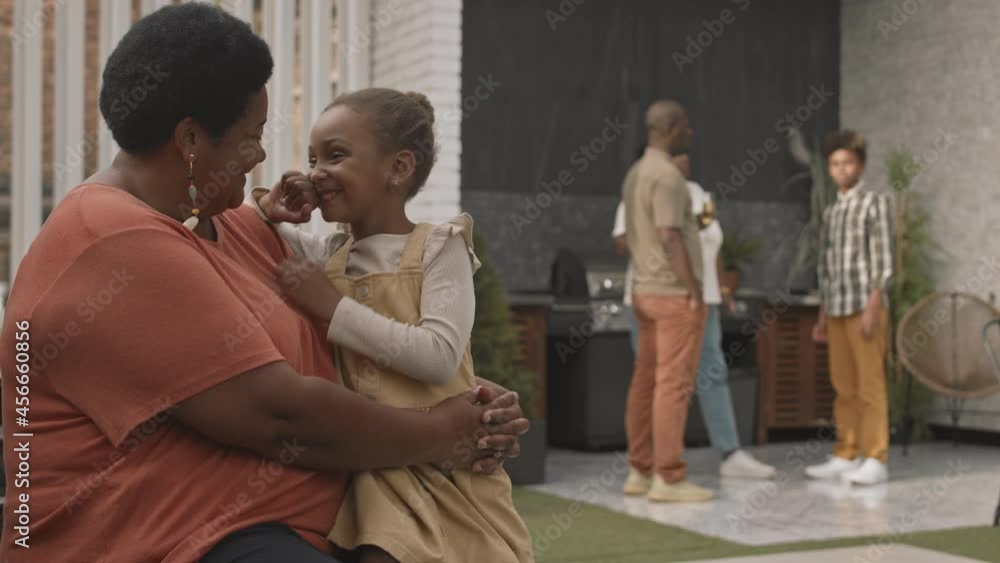 Medium long of mature African-American woman sitting with her adorable little granddaughter, talking and smiling, blurred family members standing on background Medium long of mature African-American woman sitting with her adorable little granddaughter, talking and smiling, blurred family members standing on background