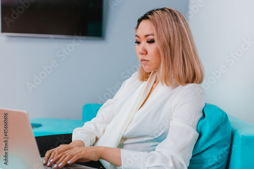 A young woman of Asian appearance with blond hair sits on a blue sofa. The girl works at home during the quarantine.