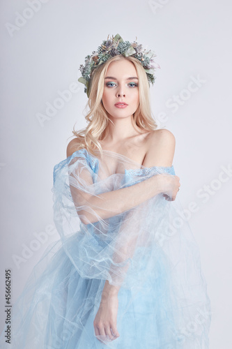Charming fairy woman in blue ethereal dress and wreath on head on white background, gentle mysterious blonde girl with perfect skin and makeup. Cleanliness, body care and skin
