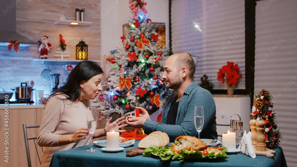 Man proposing with engagement ring to woman at festive dinner. Couple getting engaged while celebrating christmas eve with chicken meal and glasses of champagne. Cheerful people in love