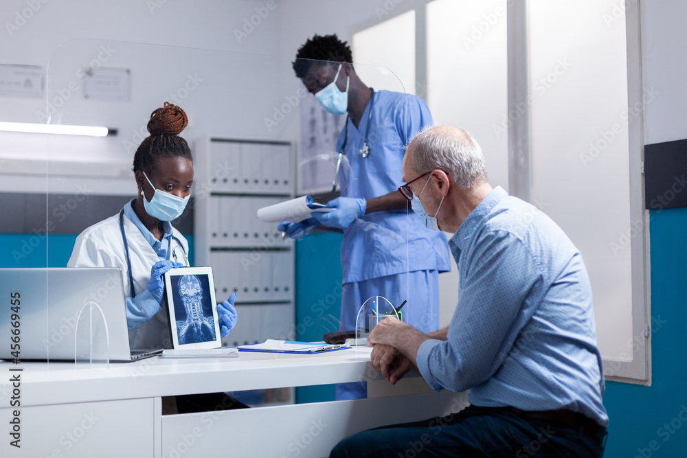 Obraz premium Black professional team of people explaining x ray to old sick man sitting at desk. African american medic and black nurse with radiography on tablet showing scan to aged patient