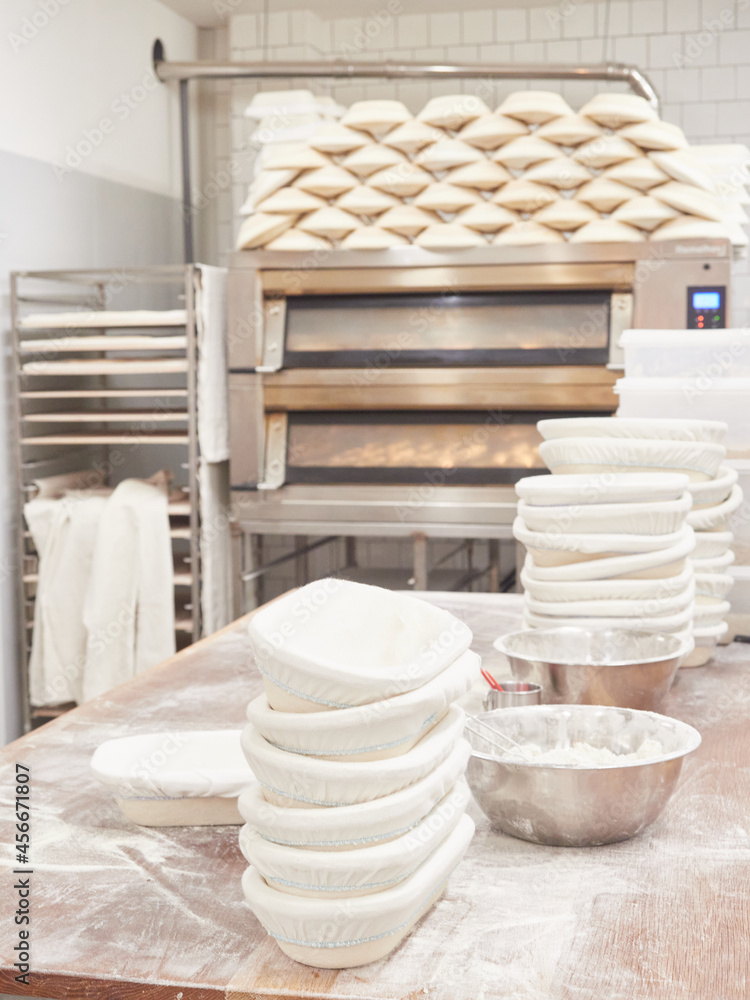 scene inside a bakery: work table for kneading bread with stacked bread pans and flour bowls, ovens and cooling rack trolley behind scene inside a bakery: work table for kneading bread with stacked bread pans and flour bowls, ovens and cooling rack trolley behind