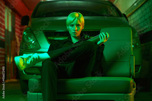 a girl with short hair sits in the garage near the car and holds a bat in her hand. Modern photo in neon green light. High quality photo