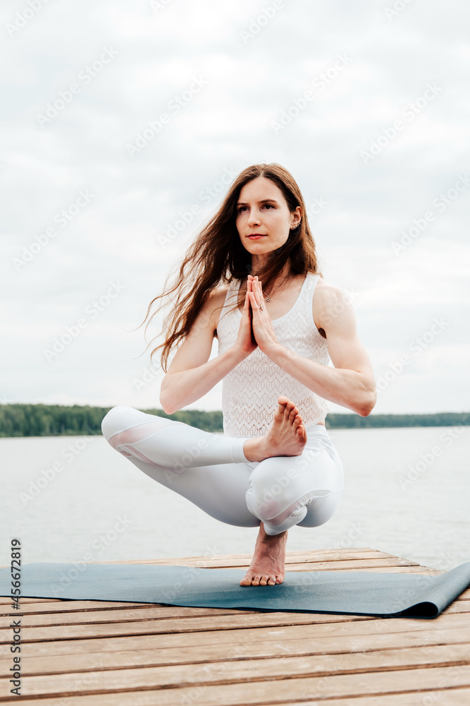 Beautiful young woman in white sports clothes is sitting on  wooden pier on lake in  balance yoga position with namaste gesture.