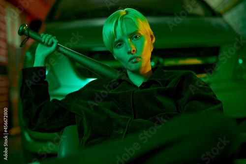fashionable shooting in neon green light girl in a tracksuit sits near the car and holds a bat behind her back. High quality photo