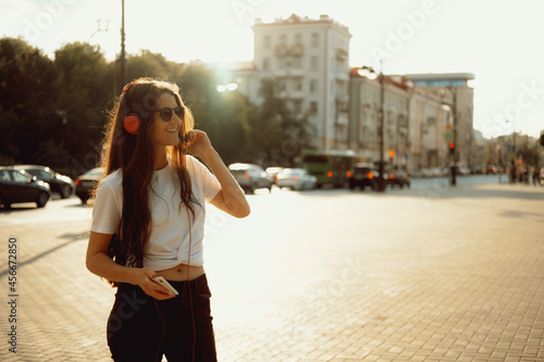 Beautiful happy young woman using cell phone and headphones and listening music in the city streets. Positive emotions, lifestyle expressive portrait in summer. © dvulikaia
