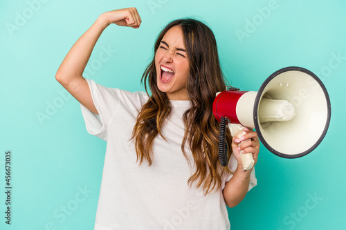 Young caucasian woman holding a megaphone isolated on blue background raising fist after a victory, winner concept.