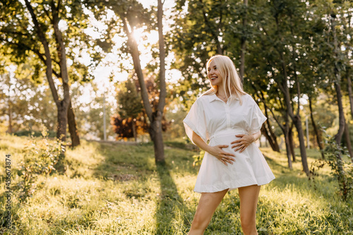 Happy pregnant woman outdoors on a sunny day, smiling, wearing a white mini dress. © Bostan Natalia