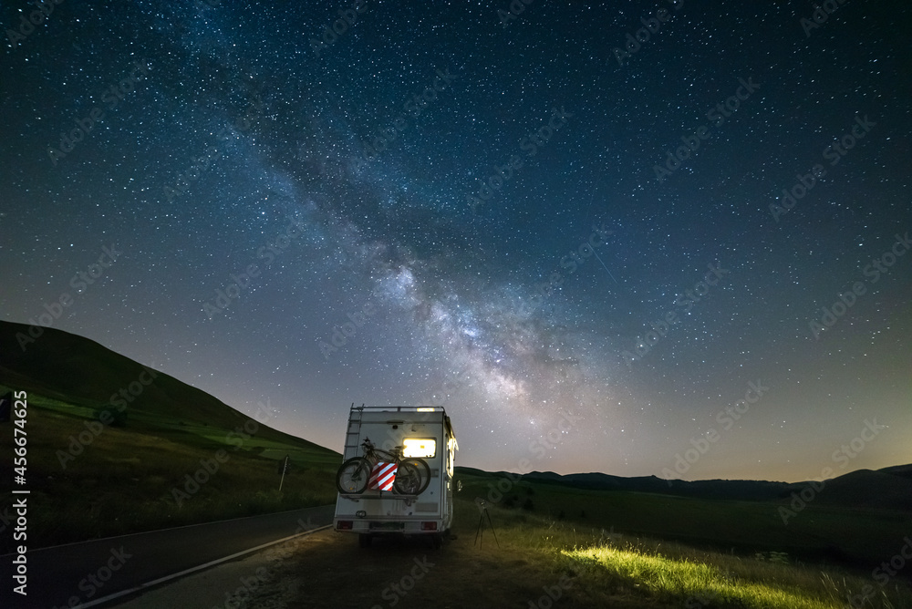 Camper van on road side in beautiful night landscape under the stars. The Milky Way galaxy arc and stars over illuminated motorhome. Camping freedom in italian Apennines.