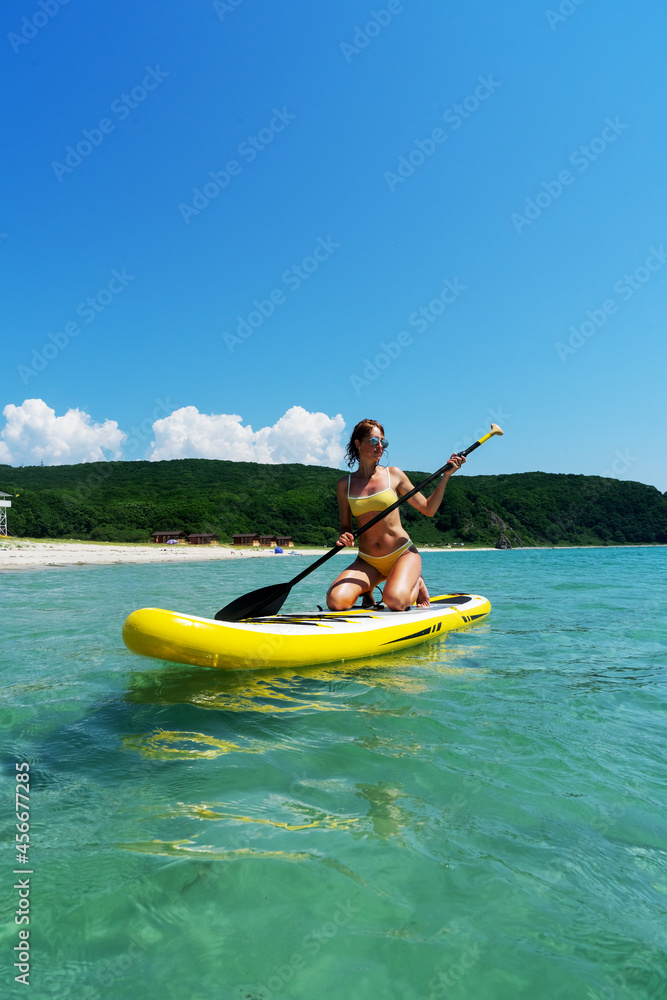 Tanned woman in bikini sitting on surfboard with paddle relaxing at sea resort enjoying summer