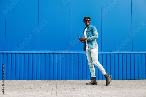 an african guy walks with a phone around the city, against the background of a blue building, makes a purchase online © Shopping King Louie