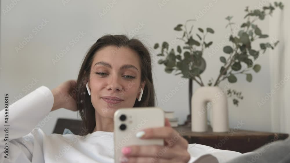 Smiling and attractive woman waving hand and talking on a video call with mobile phone in hand. Caucasian woman in white sweater chatting at living room interior Smiling and attractive woman waving hand and talking on a video call with mobile phone in hand. Caucasian woman in white sweater chatting at living room interior