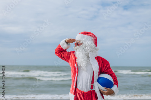 santa claus with beach ball under his arm looks at the horizon - seashore in background