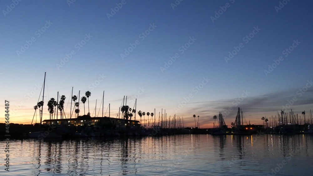 Fototapeta premium Luxury yachts sailboats floating, marina harbour quay. Sail boat masts, nautical vessels in port. Harbor fisherman village in Oceanside, California USA. Evening dusk, twilight lights and palm trees.