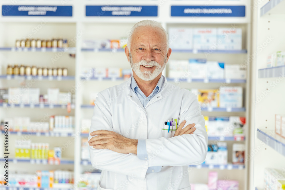 Portrait of a pharmacist in a pharmacy. Close-up of a smiling pharmacist in uniform and gray hair and beard crossed arms in the middle of the pharmacy in front of a shelf with medicines Proud worker
