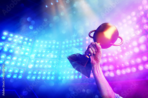 Gold trophy in winner 's hand with background spotlight on e-sport competition stage in the stadium , win concept. 